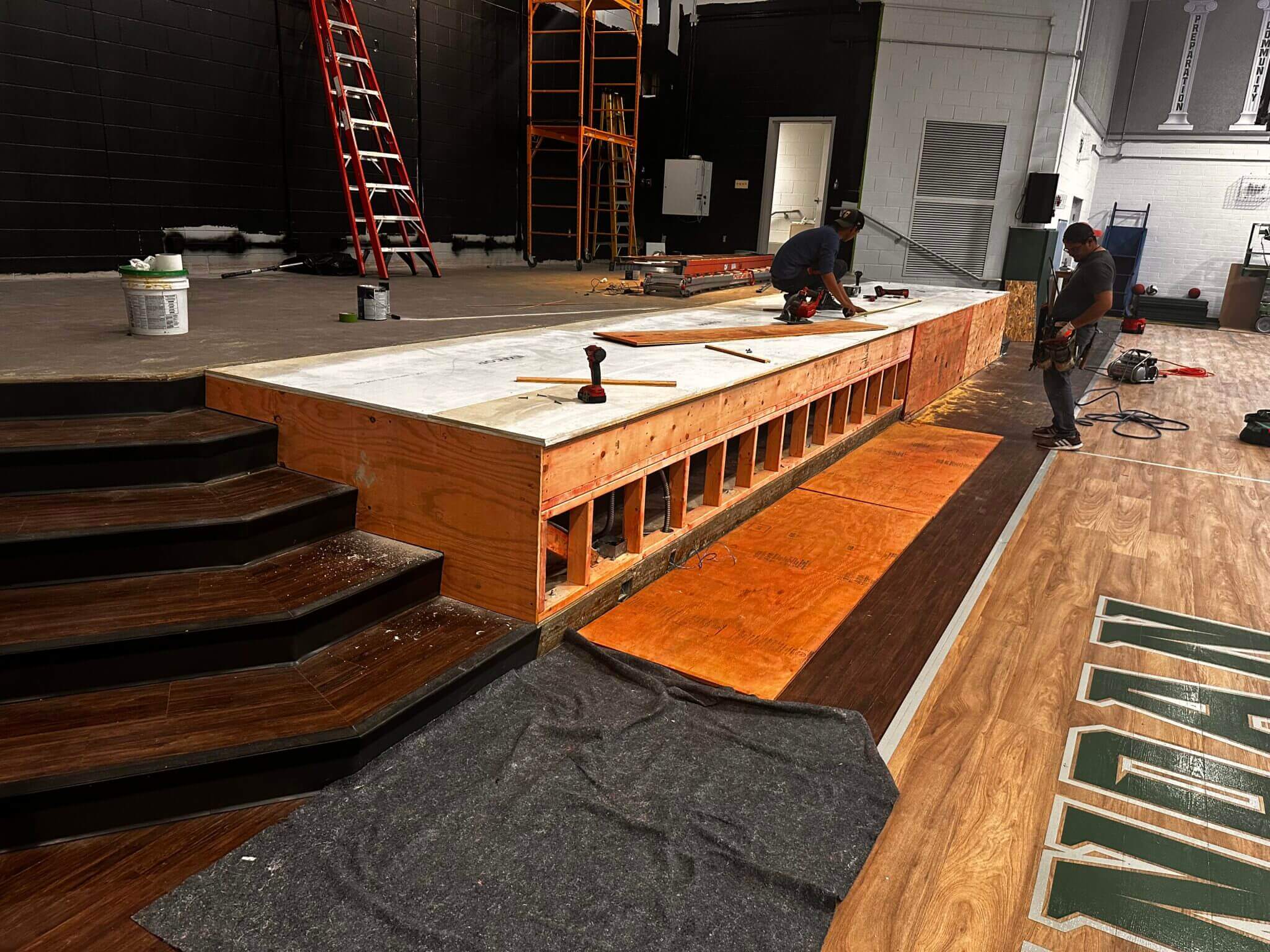 Workers building a wooden stage platform indoors.